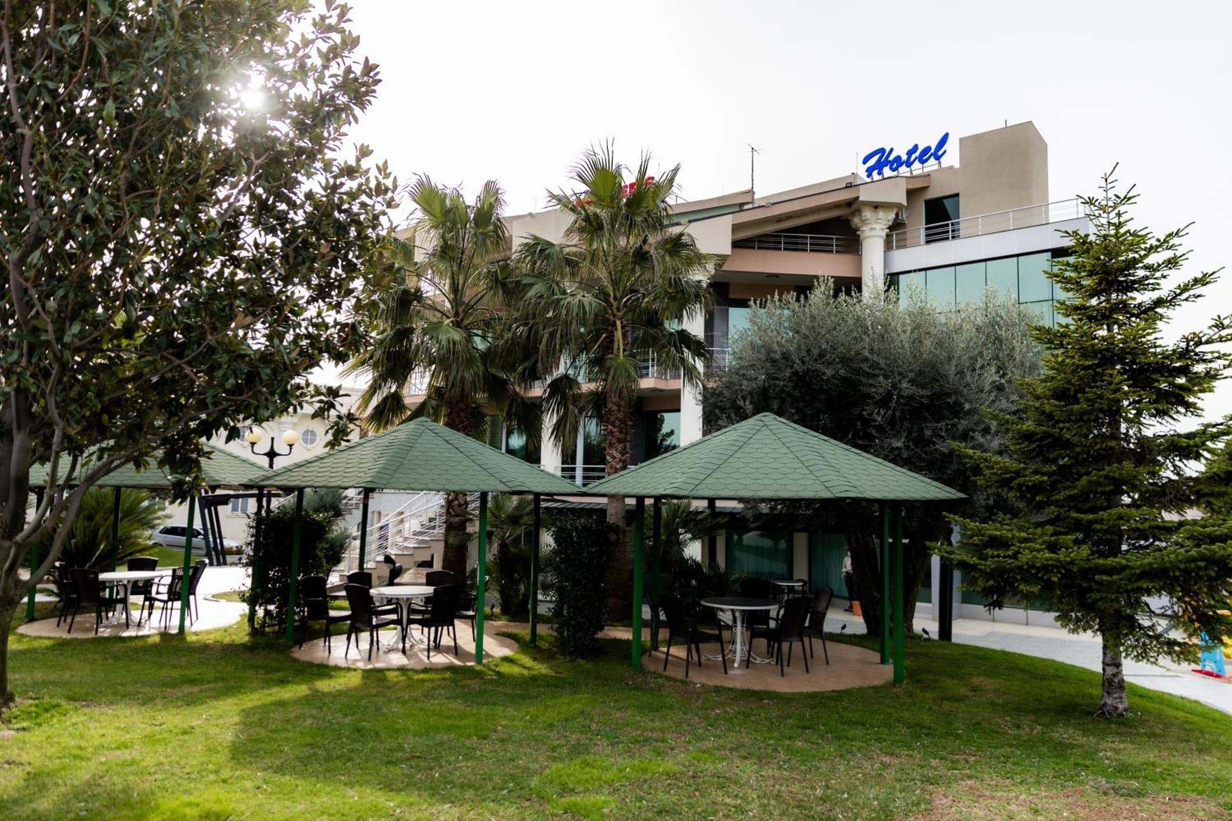 Çardakë me çati të gjelbër dhe tavolina në kopsht, me hotelin në sfond