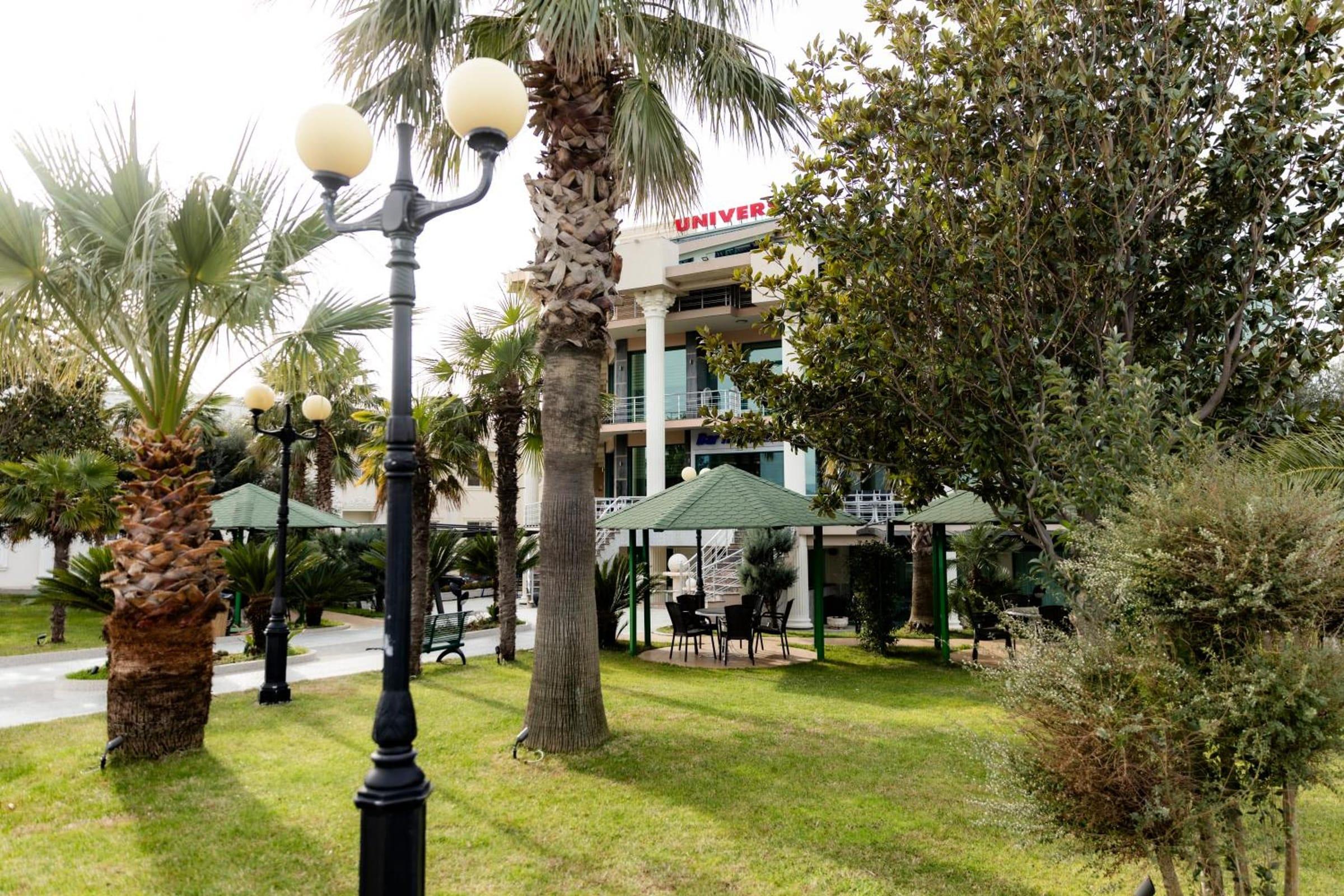 Kopshti me palma, fenerë klasikë dhe çardakë, me ndërtesën Univers pas pemëve
